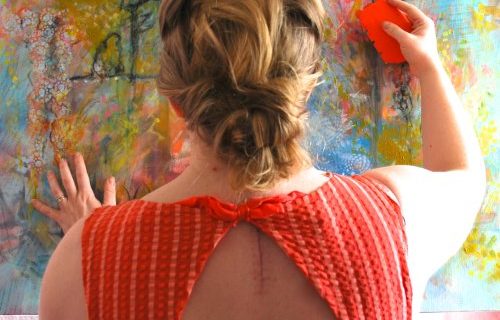 A female artist has her back to the camera, she's holding a silicone scraper up to the surface of a colourful abstract painting. her back shows scars from recent surgeries.