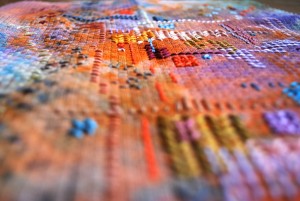 A macro photo at an angle over the texture surface of a very colourful cross stritch embroidery. The Aida fabric has been painted before it has been embroidered.
