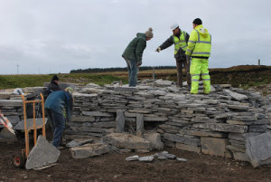 Workmen, volunteers and archaeologists dressed in high vis stand on top of the wide, low dry stone walls of a reconstructed Iron Age broch.