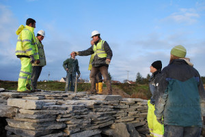 Workmen, volunteers and archaeologists dressed in high vis stand top of and around the low dry stone walls of a reconstructed Iron Age broch.