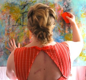 A female artist has her back to the camera, she's holding a silicone scraper up to the surface of a colourful abstract painting. her back shows scars from recent surgeries.