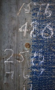 A detail photo of old weathered wood with faded blue paint in places, that has a white chalk written numbers on it.