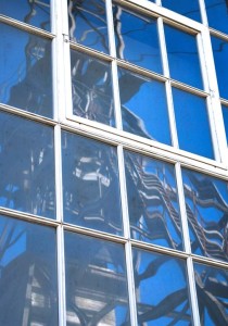 A complex detail photo showing a tall multi paned window with white painted wooden frame. In it is reflected the shape of mining machinery, all angular in shades of grey, against a pure blue sky.