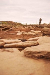 A photo showing the landscape of a working archaeological dig, showing slabs of worked sandstone strewn about, bare earth, and in the stance fencing, a spoil heap and a single figure in work clothes.