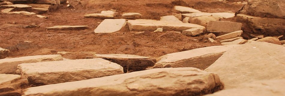 A photo showing the landscape of a working archaeological dig, showing slabs of worked sandstone strewn about, bare earth, and in the stance fencing, a spoil heap and a single figure in work clothes.