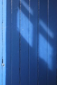 A detail photo of the painted mid blue planks of a wooden door, with shafts of light falling across them at a diagonal.