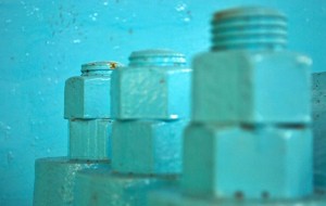 A graphic detail photo of three painted bolts, each with two large buts screwed onto them. The camera is focused on the furthest away bolt. Everything is painted in aqua green paint.