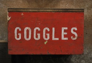 A detail photo of an old worn red painted box mounted on a wall marked 'goggles' in white painted lettering.