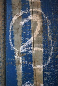 A detail photo of old weathered wood with faded blue paint in places, that has a white chalk written numbers 2 on it, enclosed in a circle.