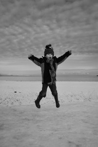 A black and whit photo of a young boy, about 5, doing a star jump against a background of a simple snowy landscape fading up with tonal similarity into a cloudy sky. He is white skinned, has his mouth open in a yell of joy, and wears all dark clothing, that contrasts to the entirely pale tones of the background.