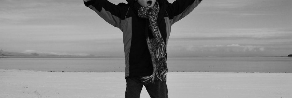 A black and whit photo of a young boy, about 5, doing a star jump against a background of a simple snowy landscape fading up with tonal similarity into a cloudy sky. He is white skinned, has his mouth open in a yell of joy, and wears all dark clothing, that contrasts to the entirely pale tones of the background.