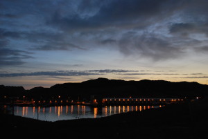 A landscape photo of low silhouetted hills under a very late sunset sky of mid and dark blue tones and wispy clouds. At the bottom of the hills are the orange evenly spaced lights of a small fishing port, which reflect into the stately blue water.