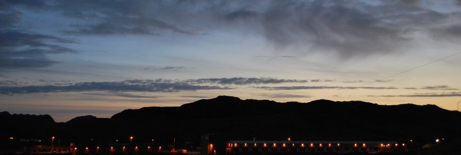 A landscape photo of low silhouetted hills under a very late sunset sky of mid and dark blue tones and wispy clouds. At the bottom of the hills are the orange evenly spaced lights of a small fishing port, which reflect into the stately blue water.