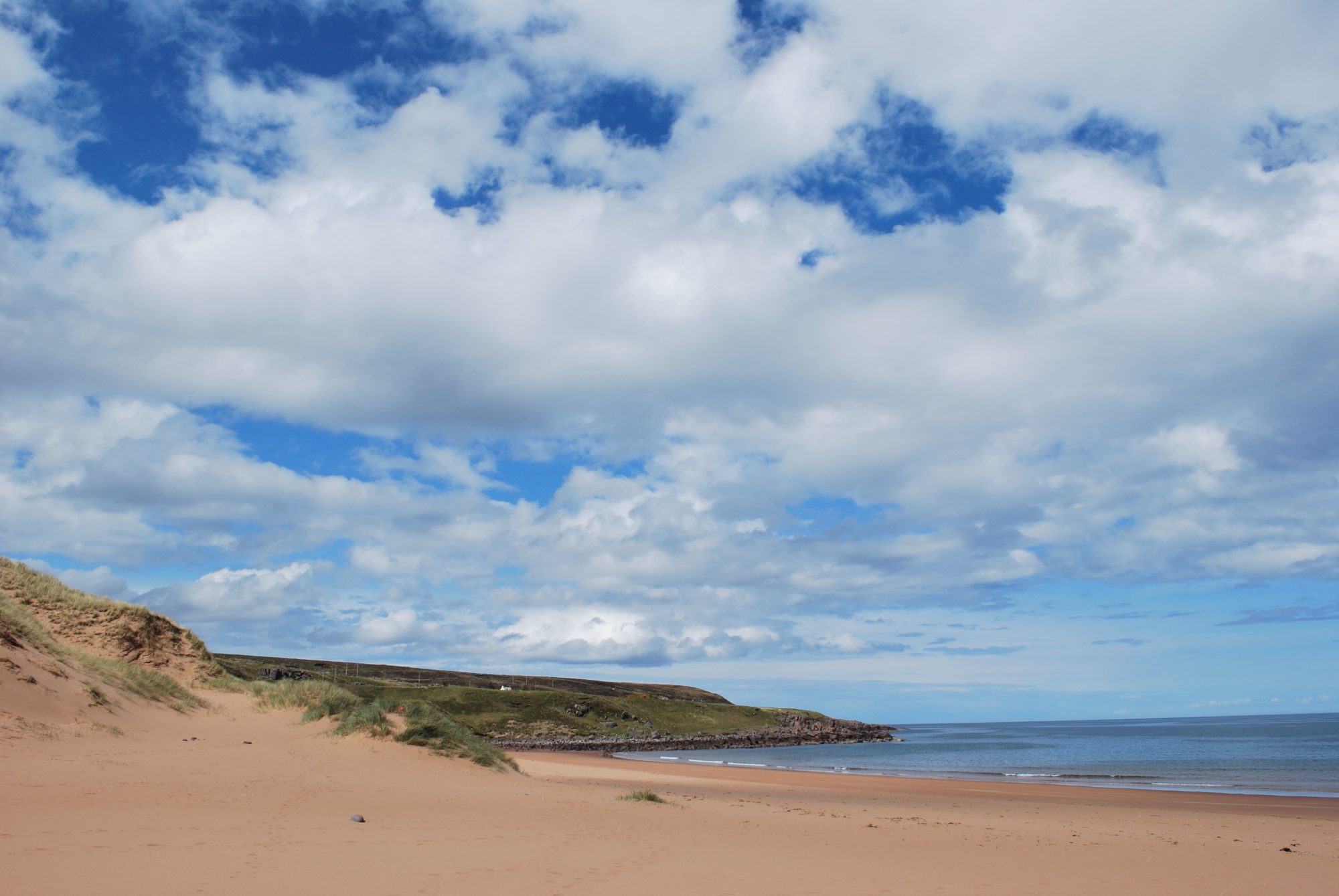 A landscape photo of buff coloured sand dunes to the left, fallen into a sandy shore which is empty of people or objects. In the distance are low grassy hills. The sky is pure vivid blue with perfect white fluffy clouds and the sea is calm.