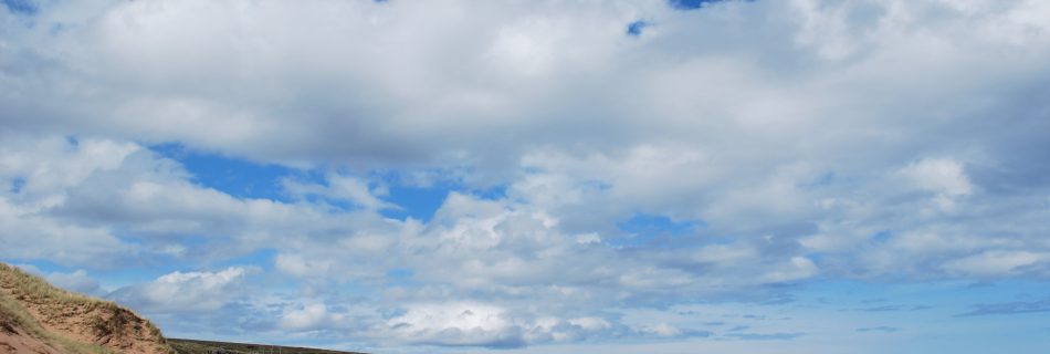 A landscape photo of buff coloured sand dunes to the left, fallen into a sandy shore which is empty of people or objects. In the distance are low grassy hills. The sky is pure vivid blue with perfect white fluffy clouds and the sea is calm.