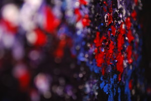 A macro photo using play with depth of field, taken across the face of an abstract painting in rich deep reds blacks and blues, showing the fine blobby texture of the paint.