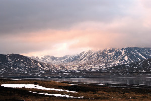A soft landscape photo of rugged grey mountains with scattered snow, fading up into hazy pink / grey snow clouds. Below a steely grey loch reflects back snow patterns.
