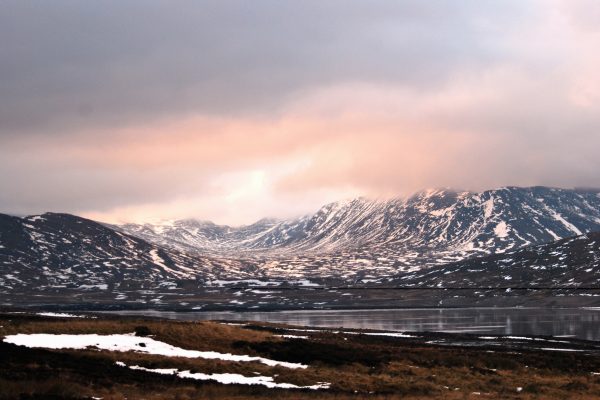 A soft landscape photo of rugged grey mountains with scattered snow, fading up into hazy pink / grey snow clouds. Below a steely grey loch reflects back snow patterns.