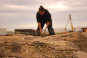 A photo of a working archaeological site. The shot clearly focuses on black of worked, bt old and distressed yellow sandstone in the foreground. Hazy, out of focus in the background can be made out a figure bend over the ground, legs apart, and the shape of a surveying apparatus on its tripod.