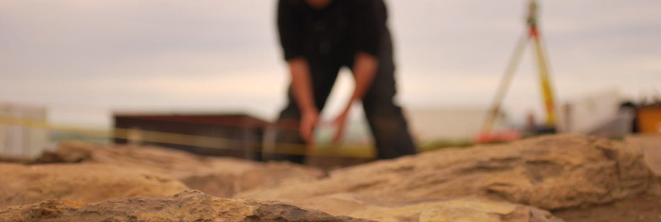 A photo of a working archaeological site. The shot clearly focuses on black of worked, bt old and distressed yellow sandstone in the foreground. Hazy, out of focus in the background can be made out a figure bend over the ground, legs apart, and the shape of a surveying apparatus on its tripod.