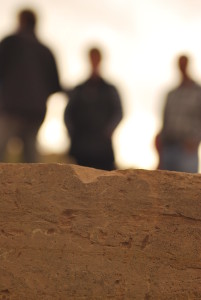 A photo showing detail of an old dressed block of chipped and distressed yellow sandstone in fine detail. In the background three figures are seen out of focus, bending into a white sky, in meeting.