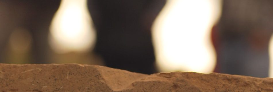 A photo showing detail of an old dressed block of chipped and distressed yellow sandstone in fine detail. In the background three figures are seen out of focus, bending into a white sky, in meeting.