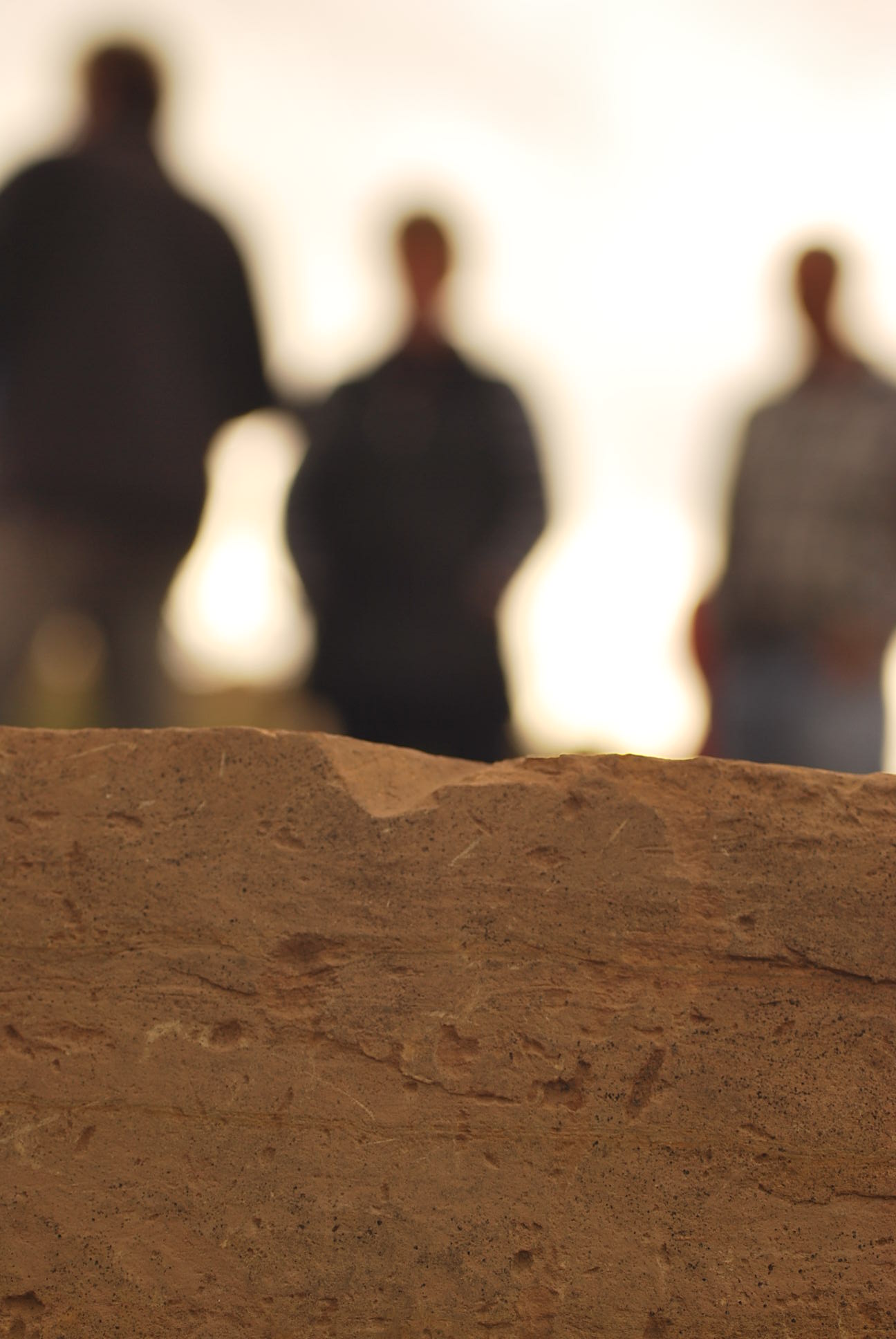 A photo showing detail of an old dressed block of chipped and distressed yellow sandstone in fine detail. In the background three figures are seen out of focus, bending into a white sky, in meeting.