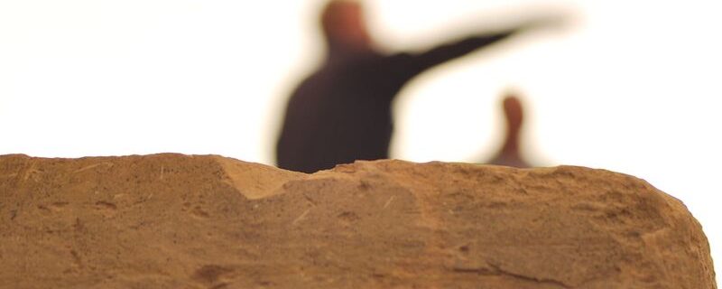 A photo of a face of old dressed sandstone in the foreground, in neat focus. It is chipped and worn. In the background two figures are out of focus against a white sky, one is pointing, gesturing a direction.