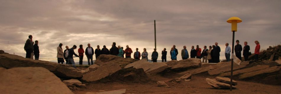 A photo at low angle cross the trenches of an archaeological dig site, treating it like a mini landscape in its won right. In the background, out of focus and silhouetted against light in the clouded grey sky are a group of around 20 people in a long continuous row, having a site meeting around the trenches far edge.