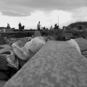 A group of distant figures in the process of work are seen from a low angle as shilouettes. They are holding clipboards, digging, or kneeling on the ground. In the foreground is a busy jumble of items. A train spotted plank lying on the ground leads the eye into the composition. Scattered rocks, flat slabs of stone and woven sandbags can be seen.