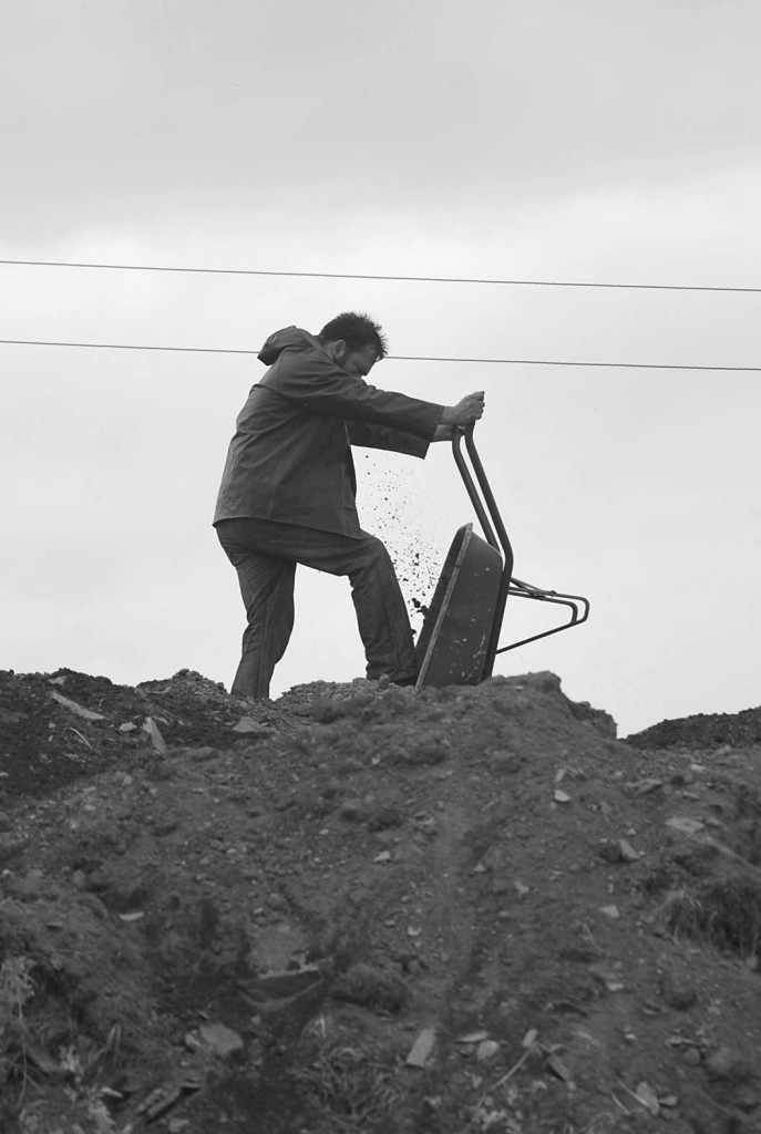 A black and white photo of a white man with shirt hair in work clothes, elevated up on a hill of earth and rubble. He is holding a wheelbarrow up and kicking it to release the earth inside onto the pile. specs of dirt are captured mid air.