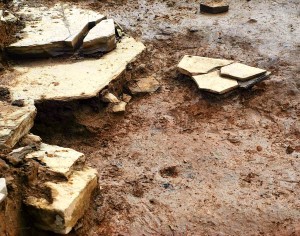A detail photo of an archaeological dig site, showing wet mud scattered with flat broken slabs of yellow sandstone.