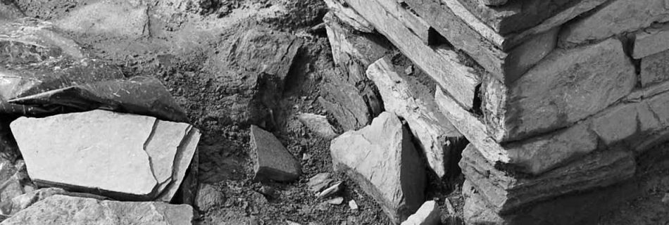 A detail photo in black and white of an archaeological dig site, showing a square pillar of stacked flat rocks rising from the dark bare earth bottom of a trench base. Black plastic sheeting and scattered rocks sit round it.
