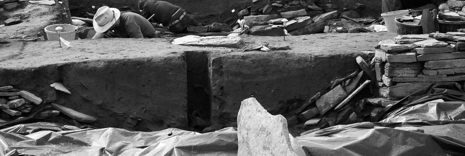 A black and white photo of a working archaeological dig site treating the trenches and walls like a landscape in its own right. Plastic sheeting covers the foreground and three bent workers can be seen behind a low earthen wall. Another wall sits behind them and beyond that flat slabs of rock stacked in undulating masses.