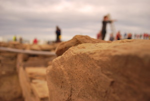 A photo of a working archaeological dig site. In the foreground are seen weathered, worked yellow sandstone rocks. In the mid and far distance are he out of focus figures of workers, standing in different places, dotter around.