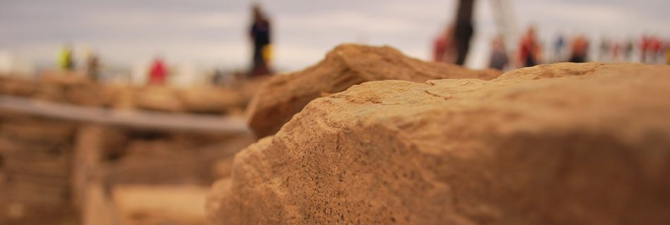 A photo of a working archaeological dig site. In the foreground are seen weathered, worked yellow sandstone rocks. In the mid and far distance are he out of focus figures of workers, standing in different places, dotter around.