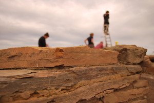 A landscape photo showing yellow flat sandstone rocks in the foreground, stacked to form a wall. In the background, distant, four figures of workers can be seen, out of focus, two stand, one bends over, the last stands on top of a set of step ladders.