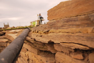 A detail photo of a working archaeological dig site, showing in the foreground right yellow sandstone slabs stacked up to form wall. A modern plastic pipe runs back to from through the middle of the photo. A standing figure can be seen in the distance holding. a clipboard and bend over it writing.