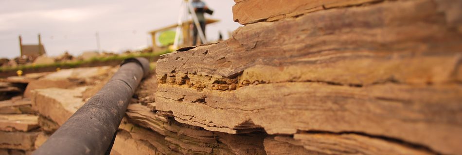 A detail photo of a working archaeological dig site, showing in the foreground right yellow sandstone slabs stacked up to form wall. A modern plastic pipe runs back to from through the middle of the photo. A standing figure can be seen in the distance holding. a clipboard and bend over it writing.