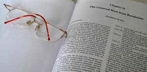 A detail photo of an open archaeology textbook with a page title 'chapter 11, the grooved ware from barn house'. a pair of delicate metal rimmed reading glasses sit on the adjacent blank page.