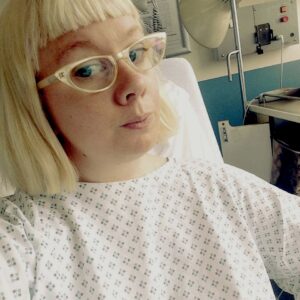 A white 30s woman with bobbed blonde hair and cat eye glasses sits in a hospital bed in an operating gown, with medical equipment in the background. 