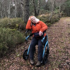 A white woman in hiking clothes sits to rest on the seat of her Rollz rollator whilst hiking in woodland. She looks exhausted.