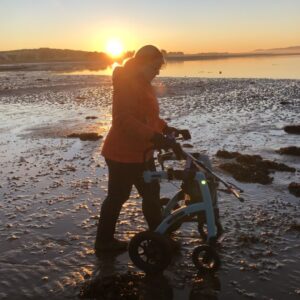 A short woman in hiking clothing and walking boots is seen in silhouette against a sunset sky, walking over rippled coastal mudflats with a rugged rollator mobility aid, with air tyres.
