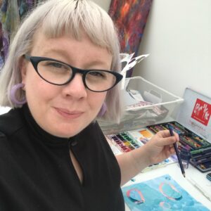 A white woman with silver blonde bobbed hair smiles at the camera. She is drawing and surrounded with art studio paraphernalia.