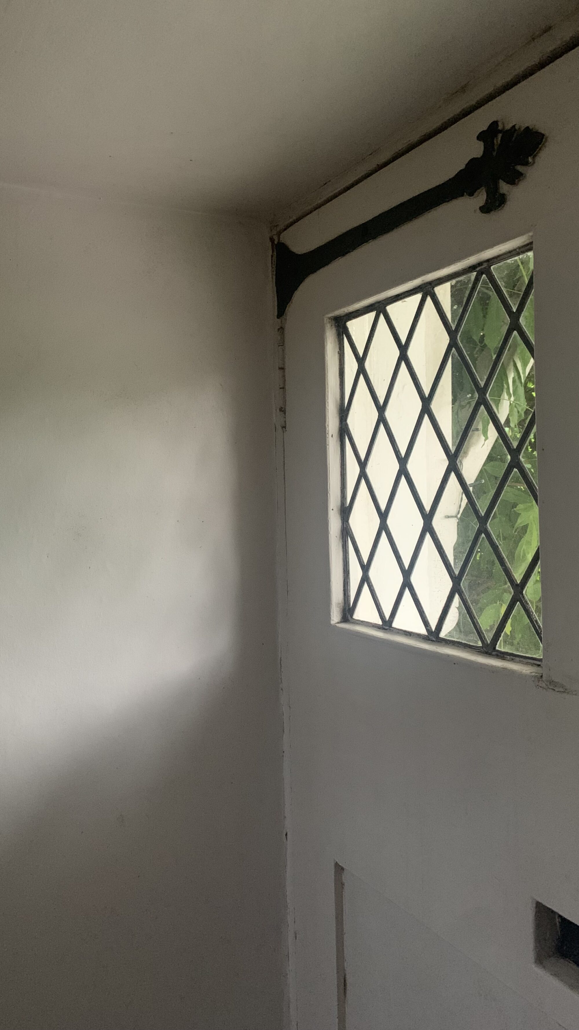 A Photo of a very old, heavy, white-painted wooden front door with lead light window to the upper part casting a shadow of light and dark pattern onto a plain white internal wall.