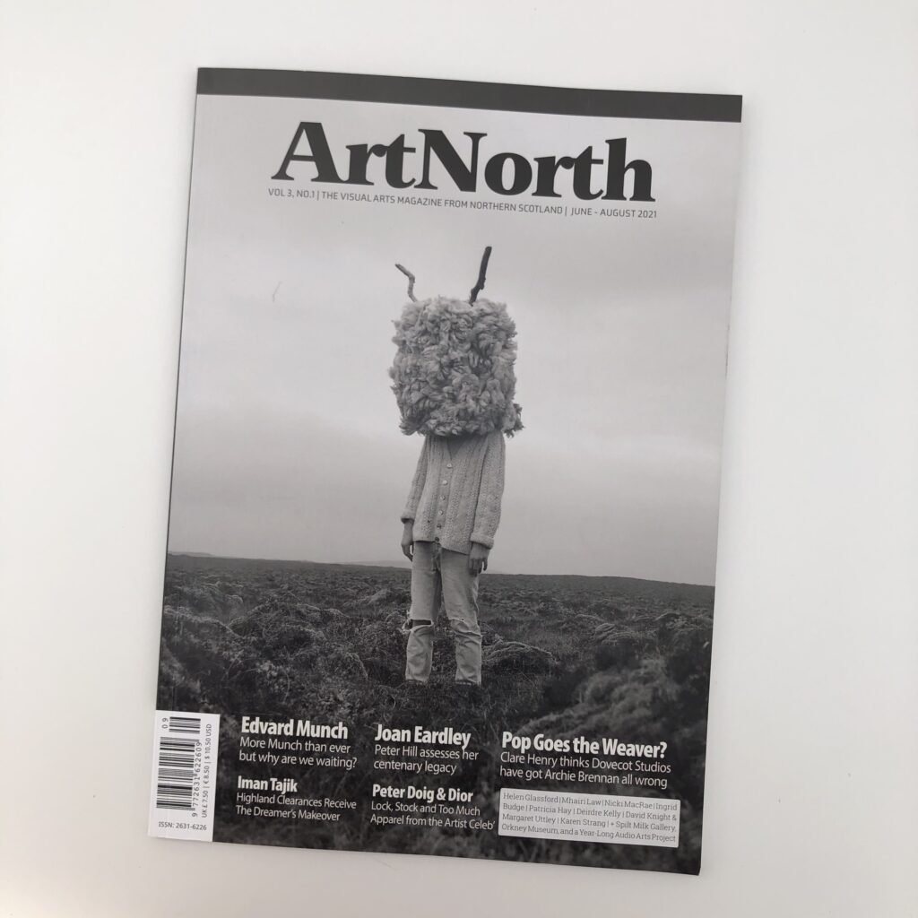 A photo of 'Art North' magazine cover on white desk. The black and white design features a figure wearing furry horned mask in a desolate grassy landscape.